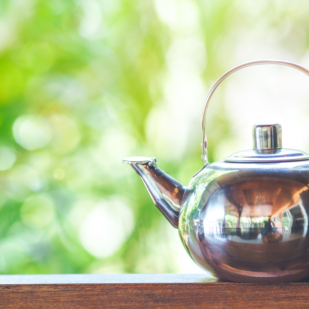 Tea kettle on a wooden ledge with green foliage in the background.